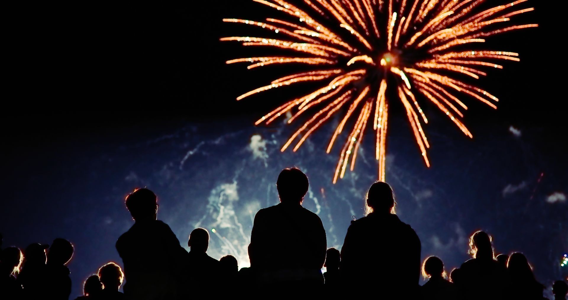 Crowd silhouetted at night watching orange fireworks burst over a glowing sky