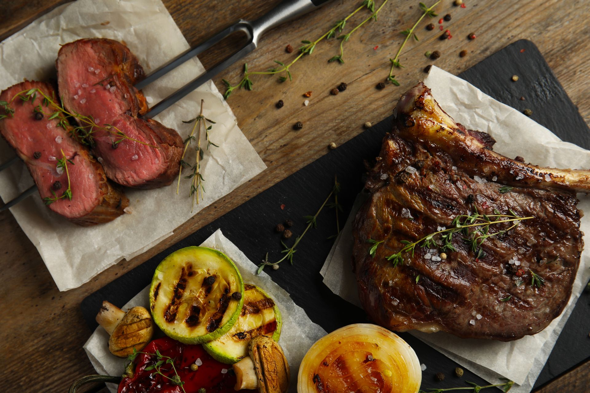 Grilled steak and sliced beef with charred vegetables on parchment and a wooden board
