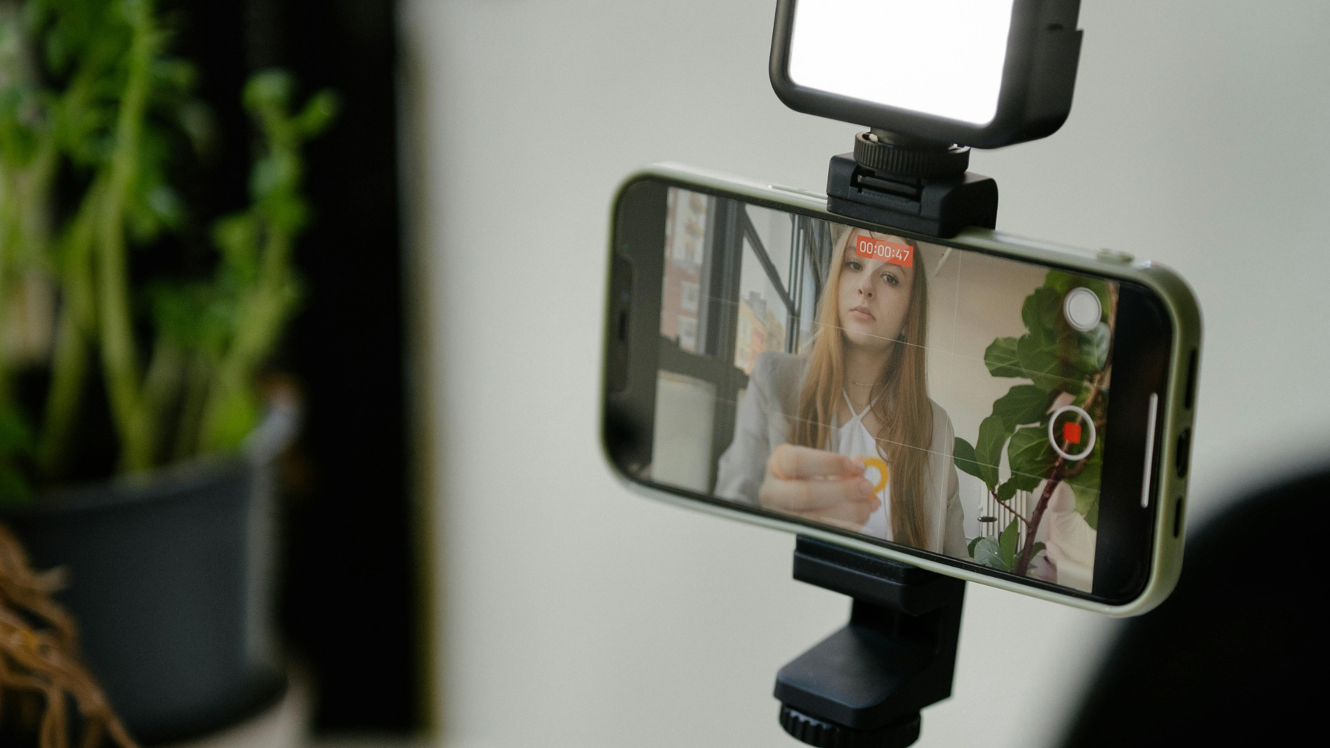 Smartphone on a tripod recording a woman speaking indoors, with a ring light above the phone.