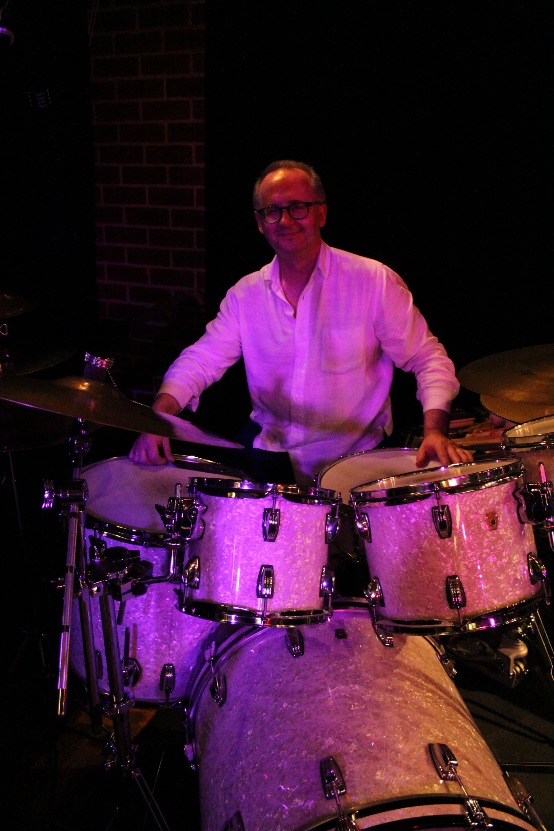A man is playing drums in a dark room with purple lights.