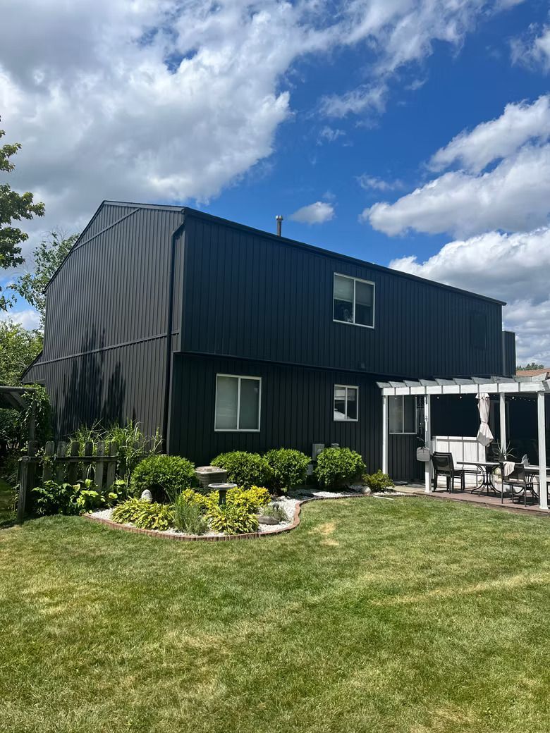 Dark, two-story house with a metal roof and siding. White window frames, lush green yard, and blue sky.