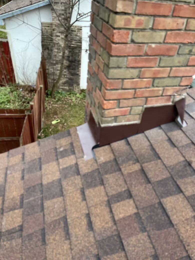 Brick chimney on a shingled roof with metal flashing; brown, beige, and green tones.