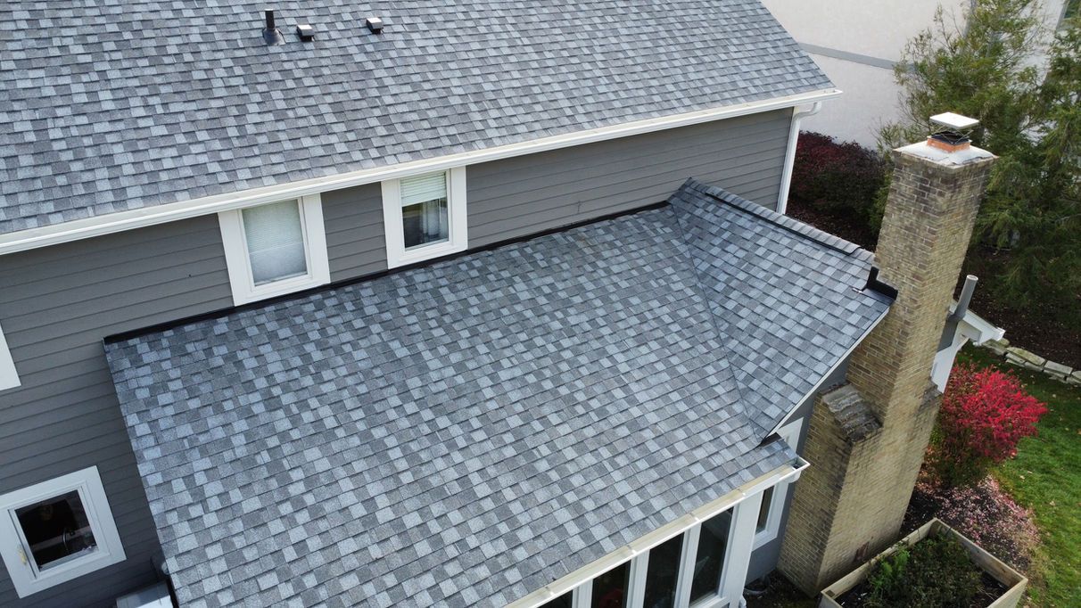 Gray shingle roof on a two-story house with a brick chimney and white window frames.