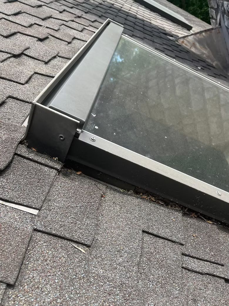 Skylight on a shingled roof, with gray metal frame. The glass is slightly dirty, angled towards the sky.