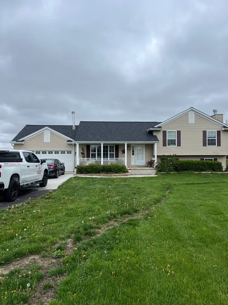A house with beige siding, a dark roof, and a green lawn. There are vehicles parked in the driveway.