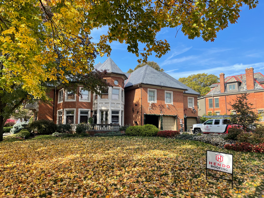 Large brick house with slate roof, front lawn covered in fallen leaves, fall foliage in the background.