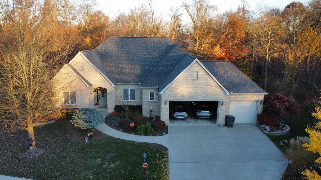 Beige brick house with attached garage, surrounded by trees. Cars inside the garage.