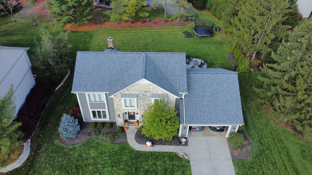 Aerial view of a two-story house with a gray roof and driveway, surrounded by green grass and trees.