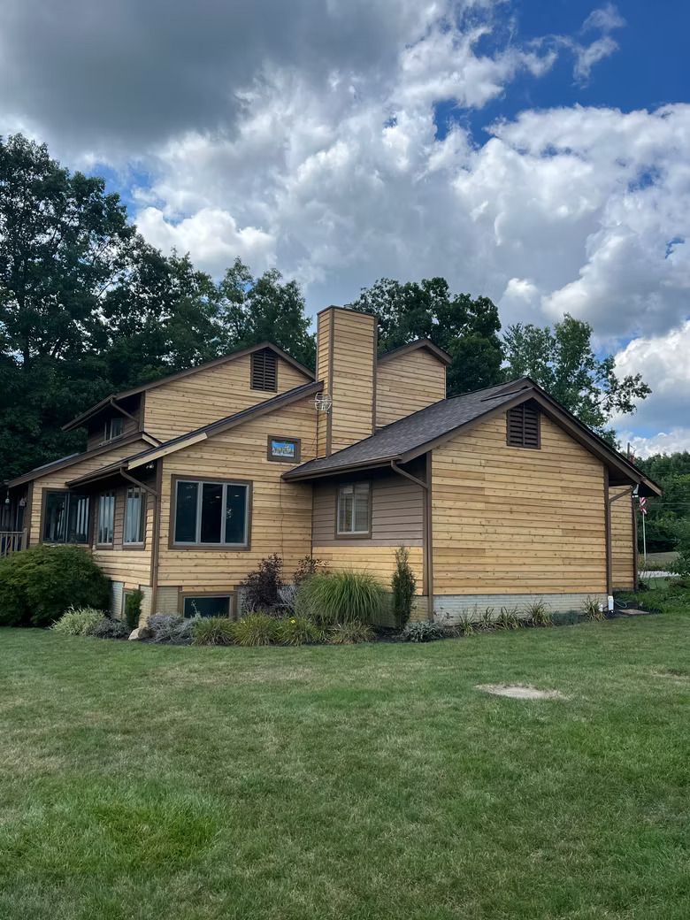 Two-story house with wood siding, brown roof, and chimney on a grassy lawn under a cloudy sky.