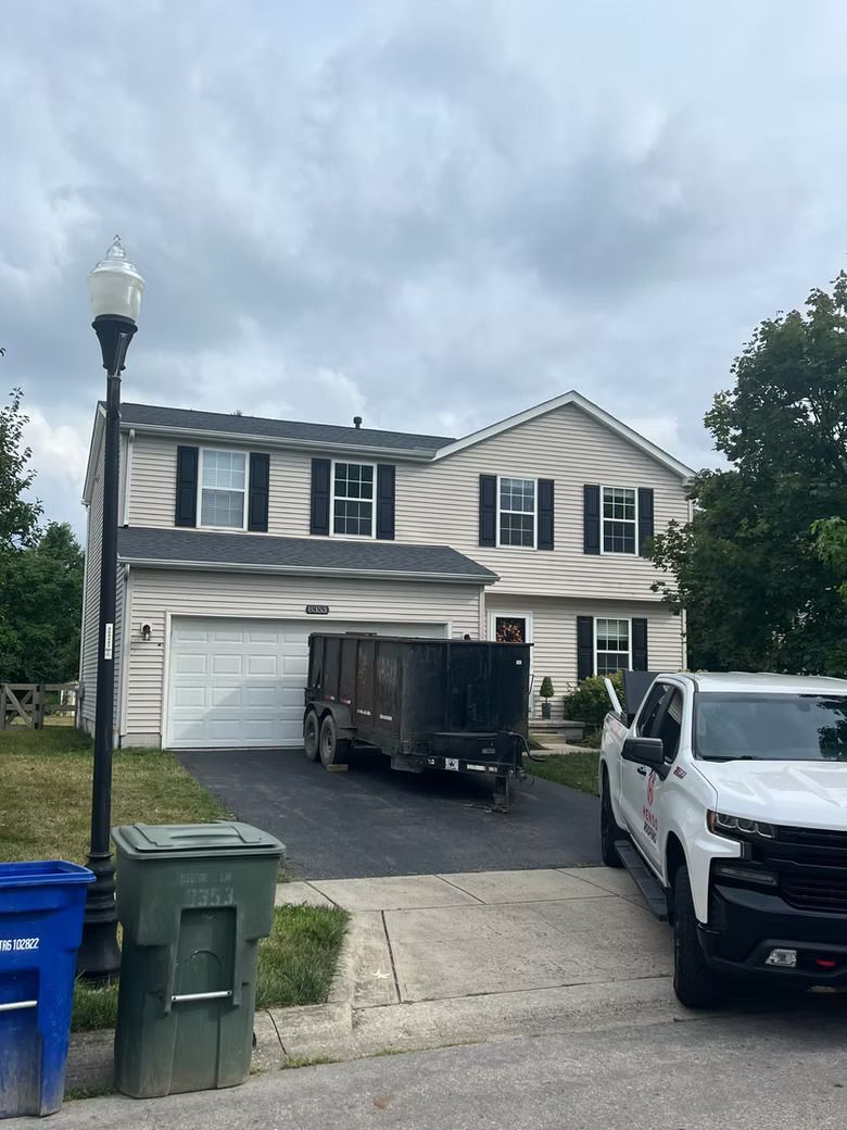 Two-story beige house with black shutters, a driveway with a dumpster, a white truck, and a lamppost.