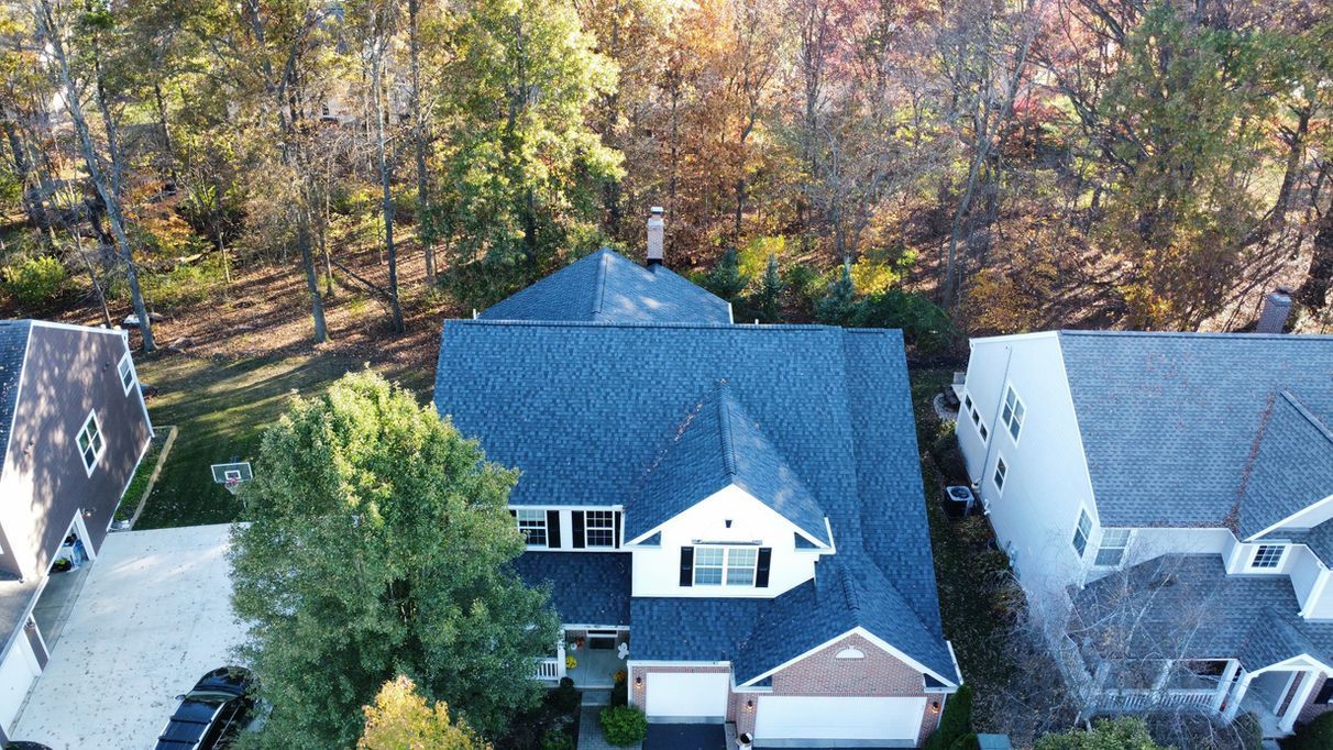 Aerial view of a two-story house with a blue roof, trees in the background, and a driveway in front.