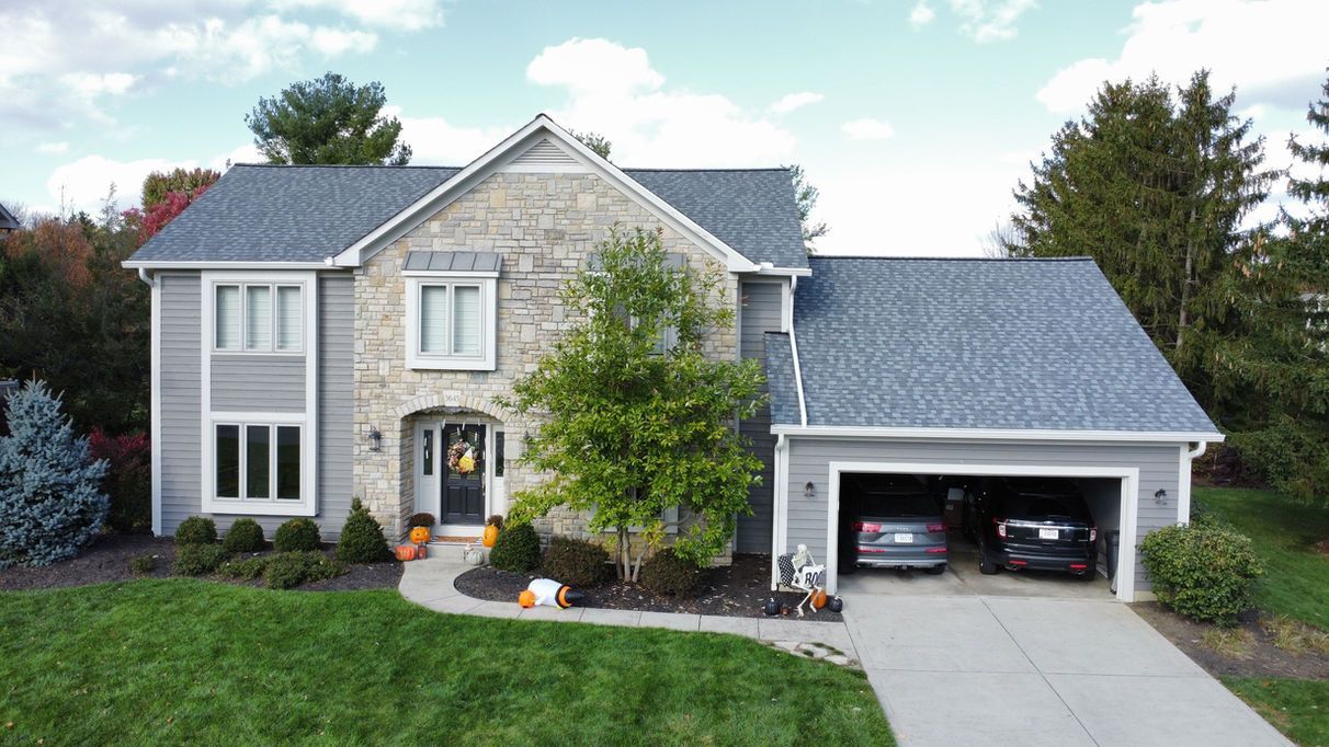 Two-story house with gray siding, stone facade, and attached garage. Cars inside. Green lawn, blue sky.