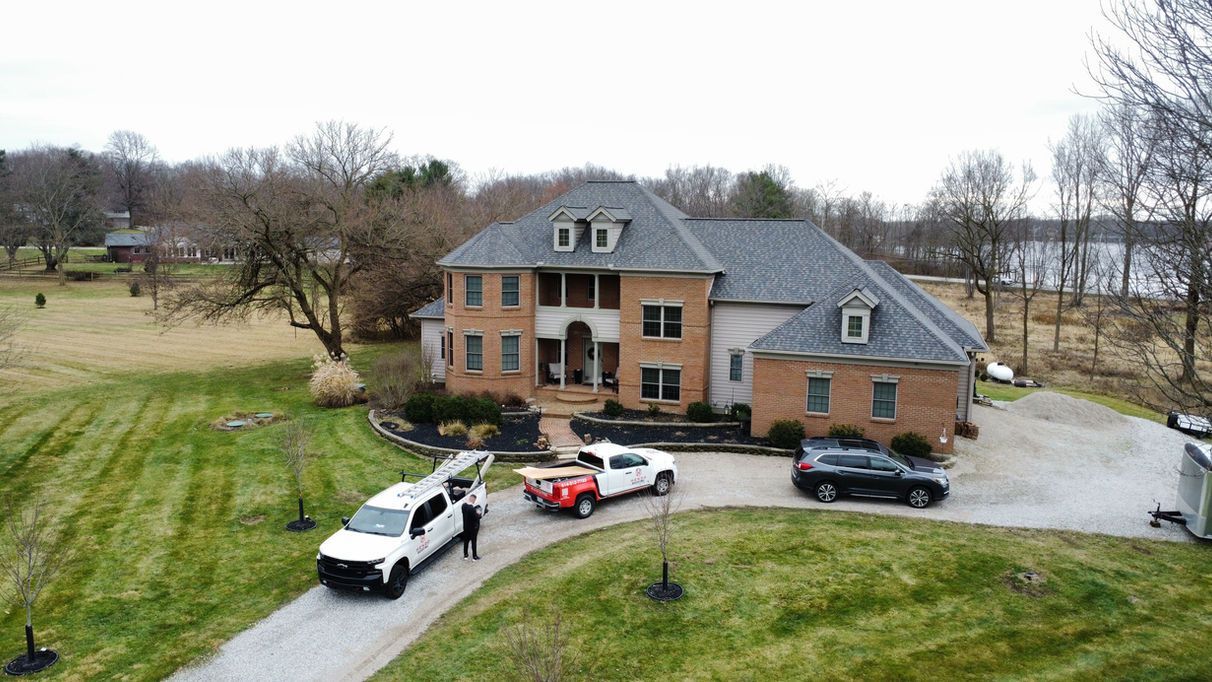 Large brick house with multiple vehicles parked in front, trees, and lake visible.