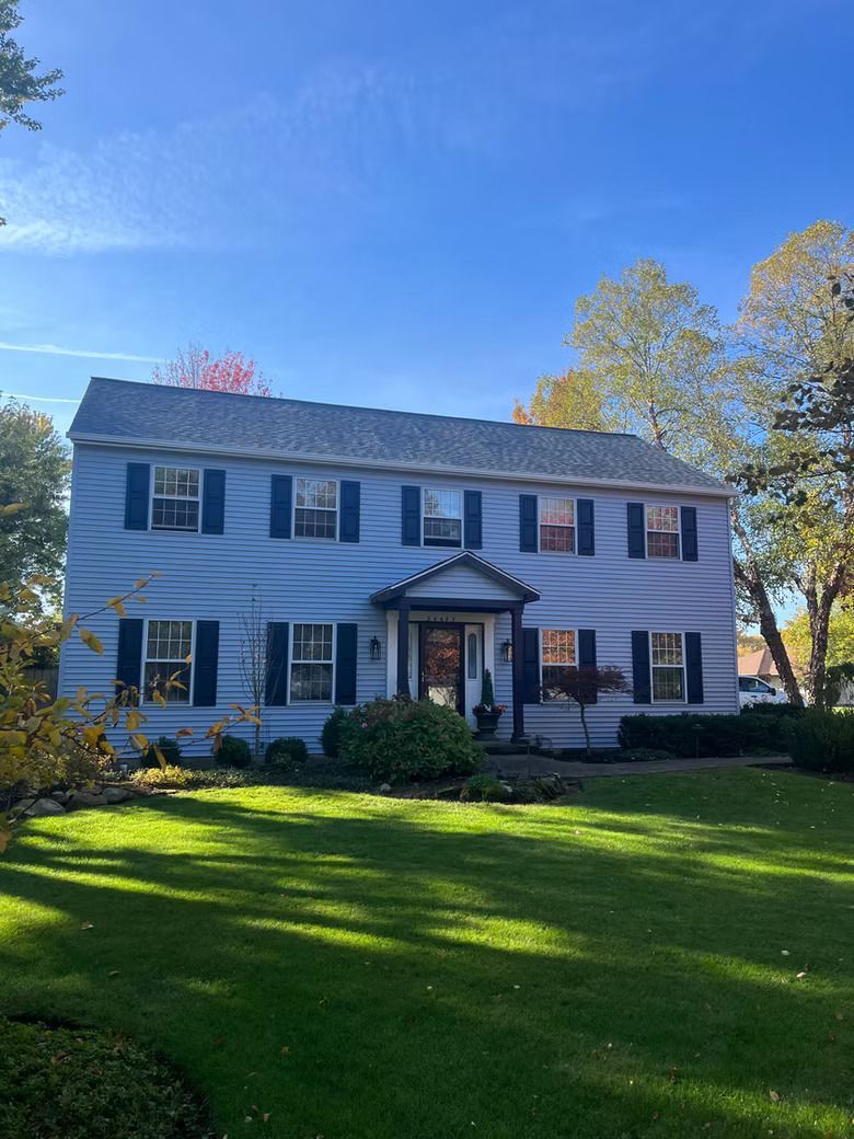 Two-story blue house with black shutters, on a green lawn with trees, under a blue sky.