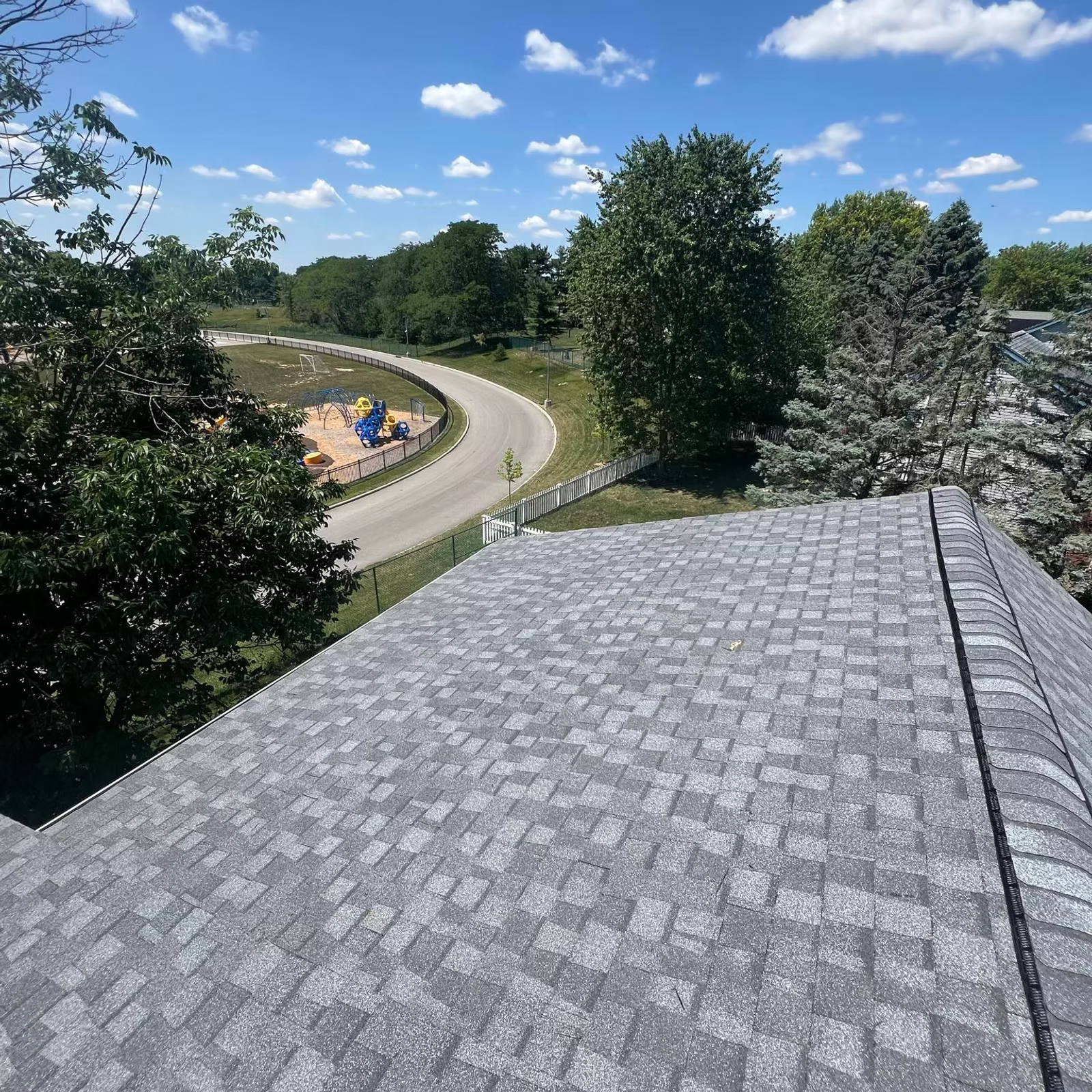 Overhead view of a gray shingled roof with a road and trees in the background under a blue sky.