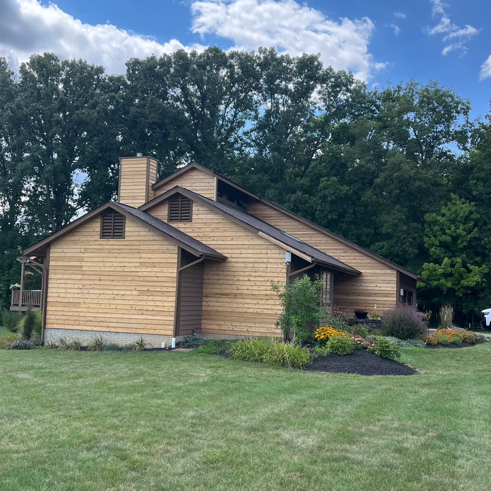 Wooden house with brown siding, chimney, and angled roof, in a grassy yard, surrounded by trees.