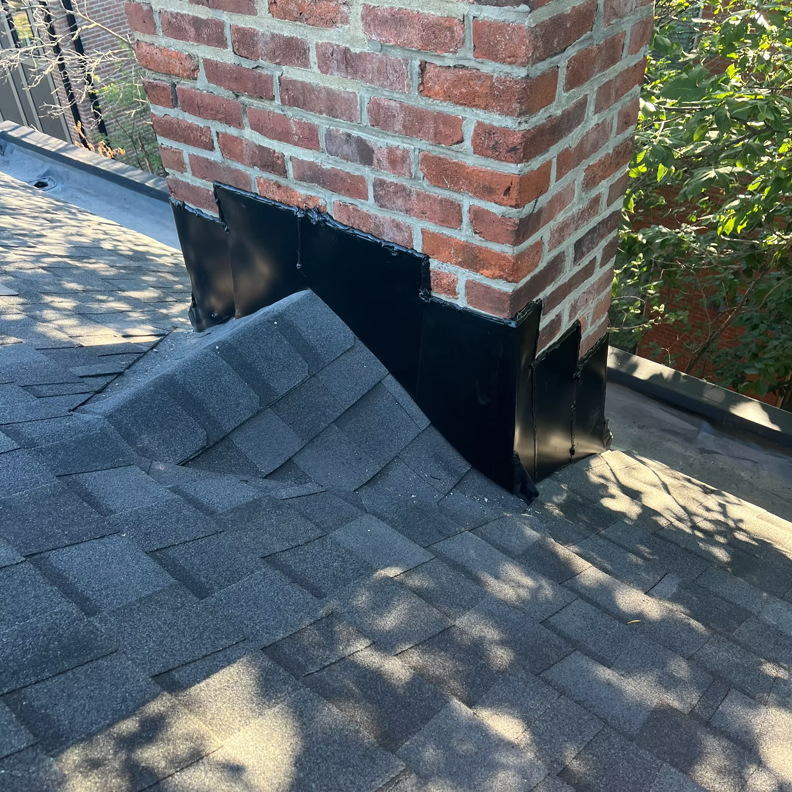 Chimney on a gray shingle roof, black flashing at the base. Brick chimney. Sunny outdoor setting.