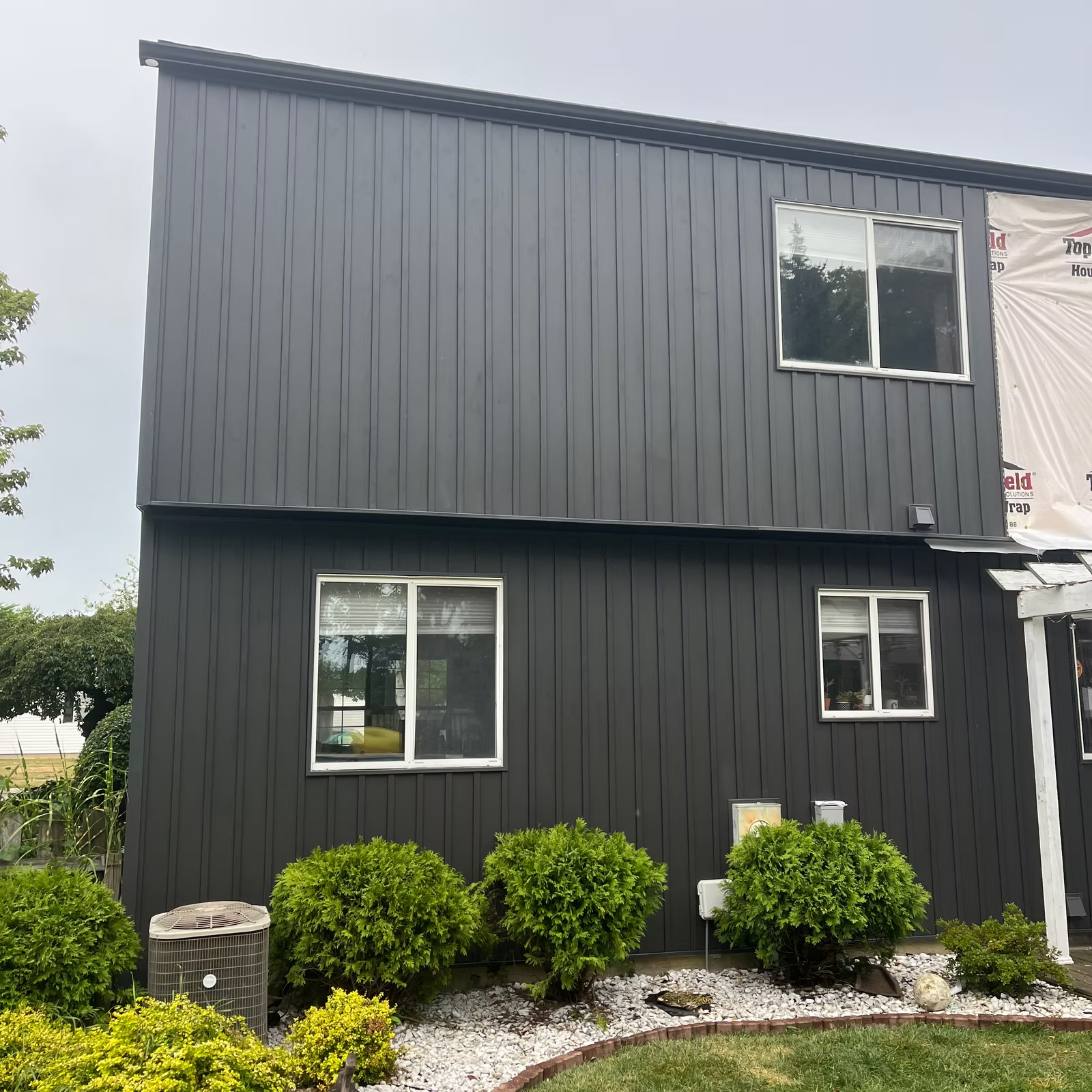 Dark gray siding on a two-story house with white-framed windows, surrounded by green shrubs and white stones.