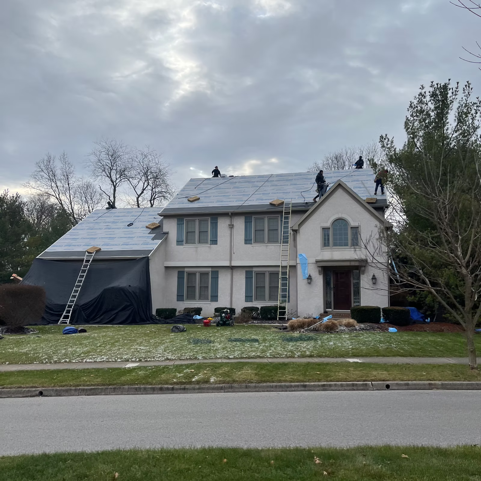 Workers on a two-story house roof, partially covered, removing old roofing. Cloudy sky, green lawn.