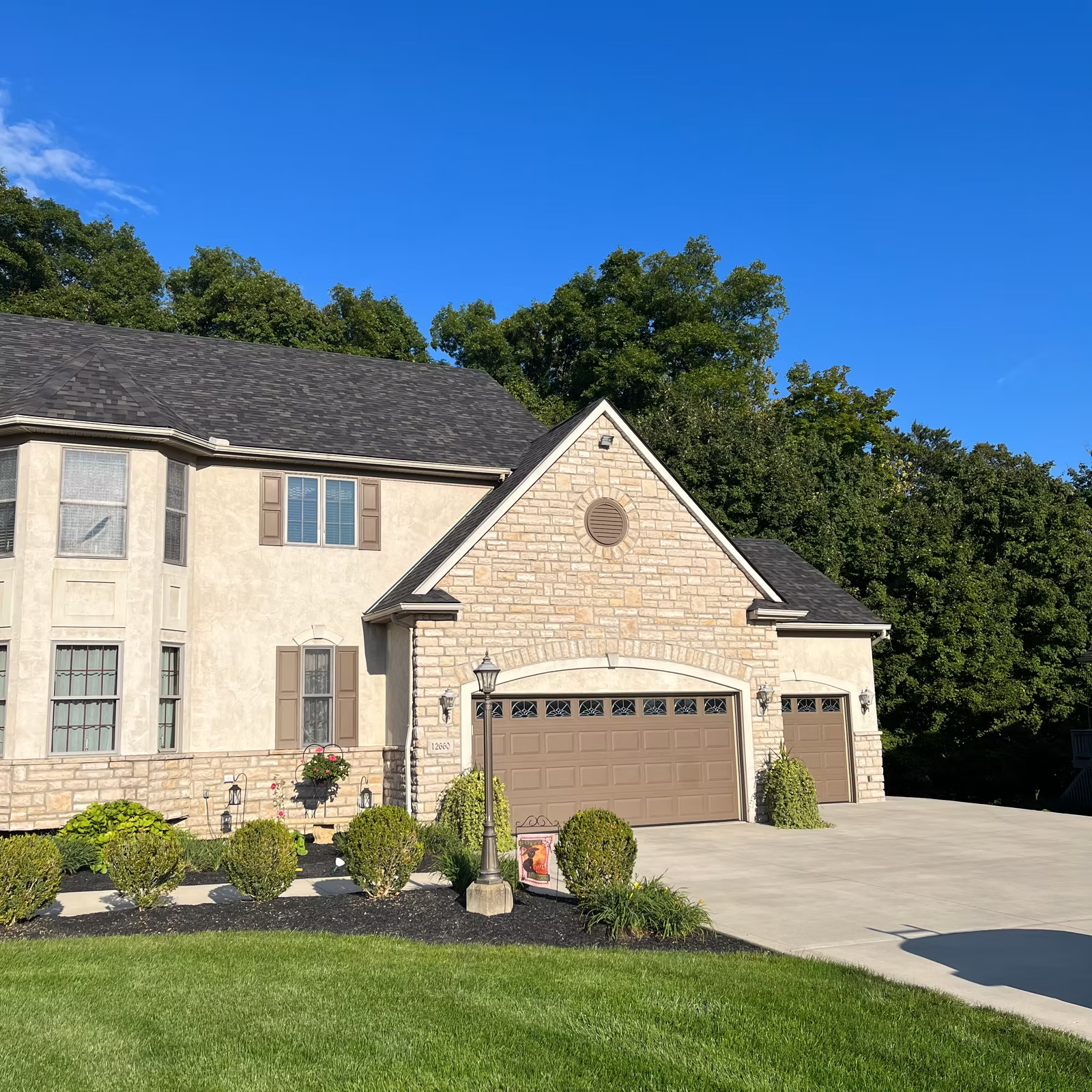 Two-story house with beige stucco and stone exterior, brown garage doors, and lush landscaping on a sunny day.