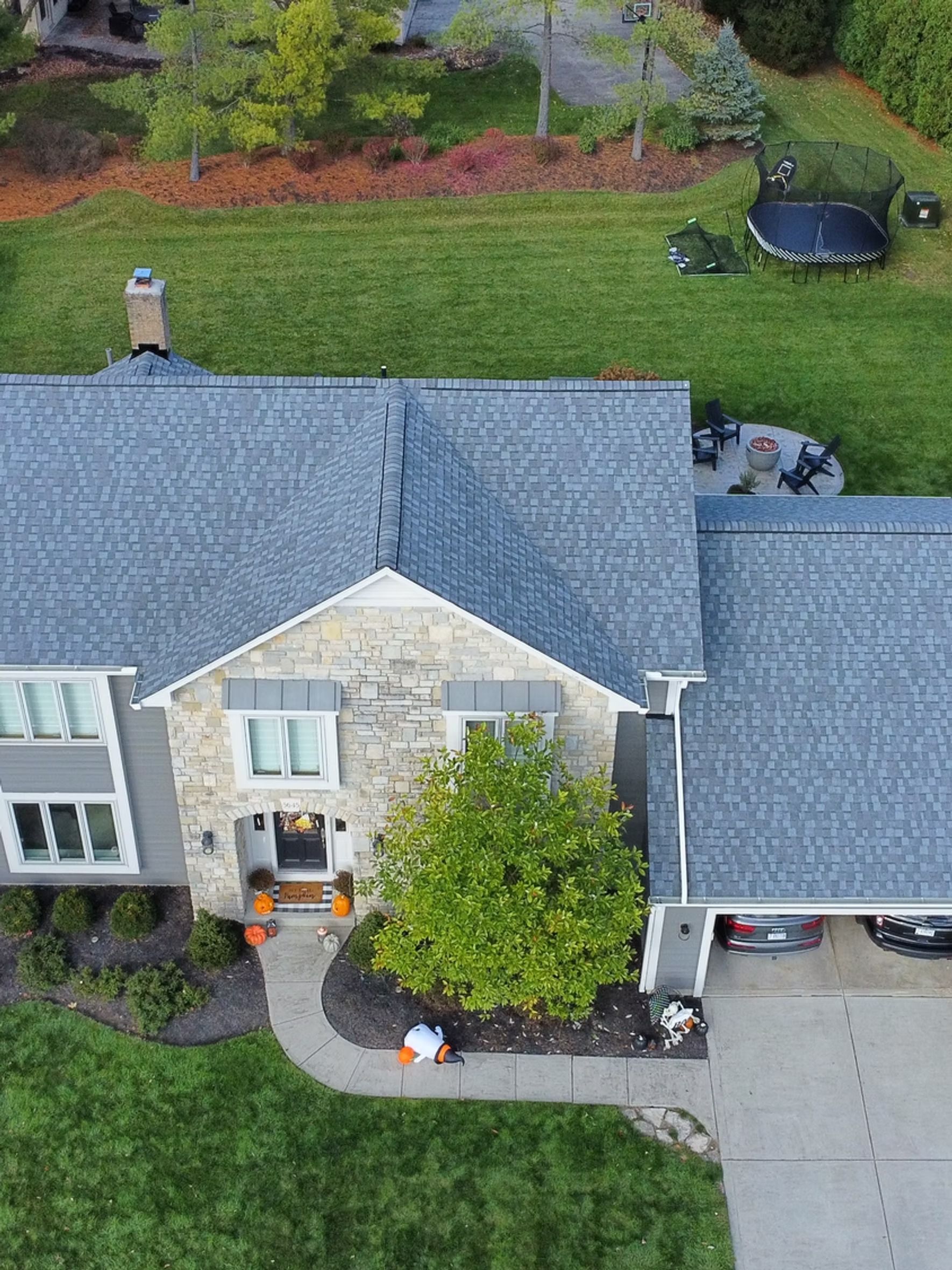 Aerial view of a gray-roofed house with stone facade, a driveway with cars, and green lawn.