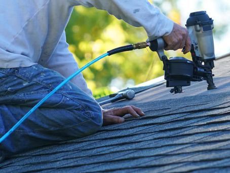 Person using a nail gun to install asphalt shingles on a roof. Blue air hose visible.