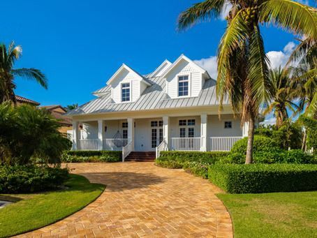 White house with dormers, porch, and brick driveway under a blue sky, surrounded by palm trees and manicured hedges.