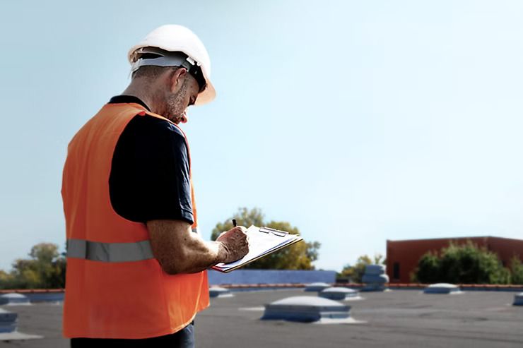 Construction worker in orange vest and hard hat, inspecting a rooftop, taking notes on a clipboard under a blue sky.