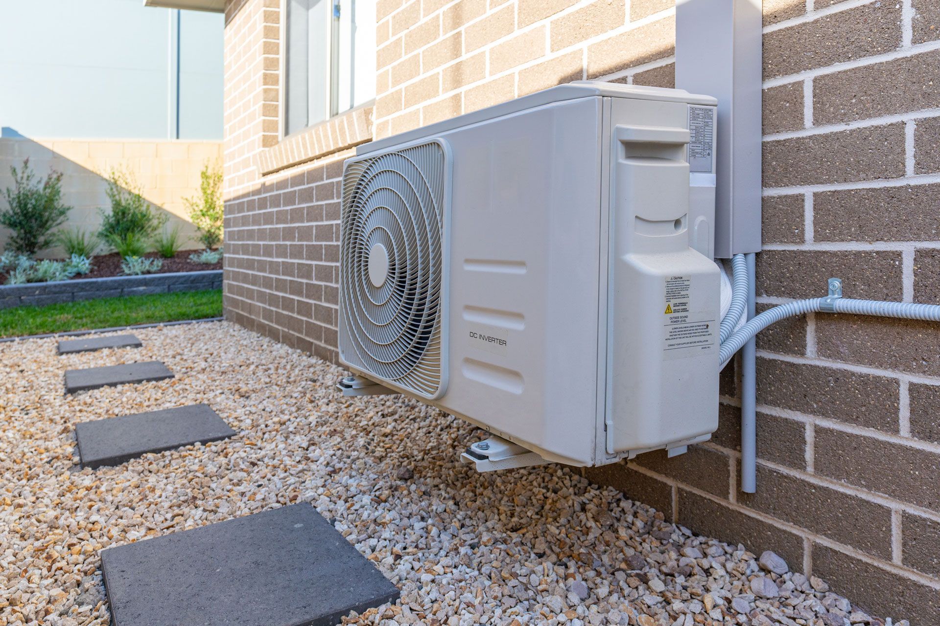 Air conditioning unit mounted on a brick wall, surrounded by gravel and stepping stones.