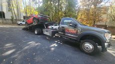 Tow truck carrying a red vehicle; parked on an asphalt driveway near trees and a building.