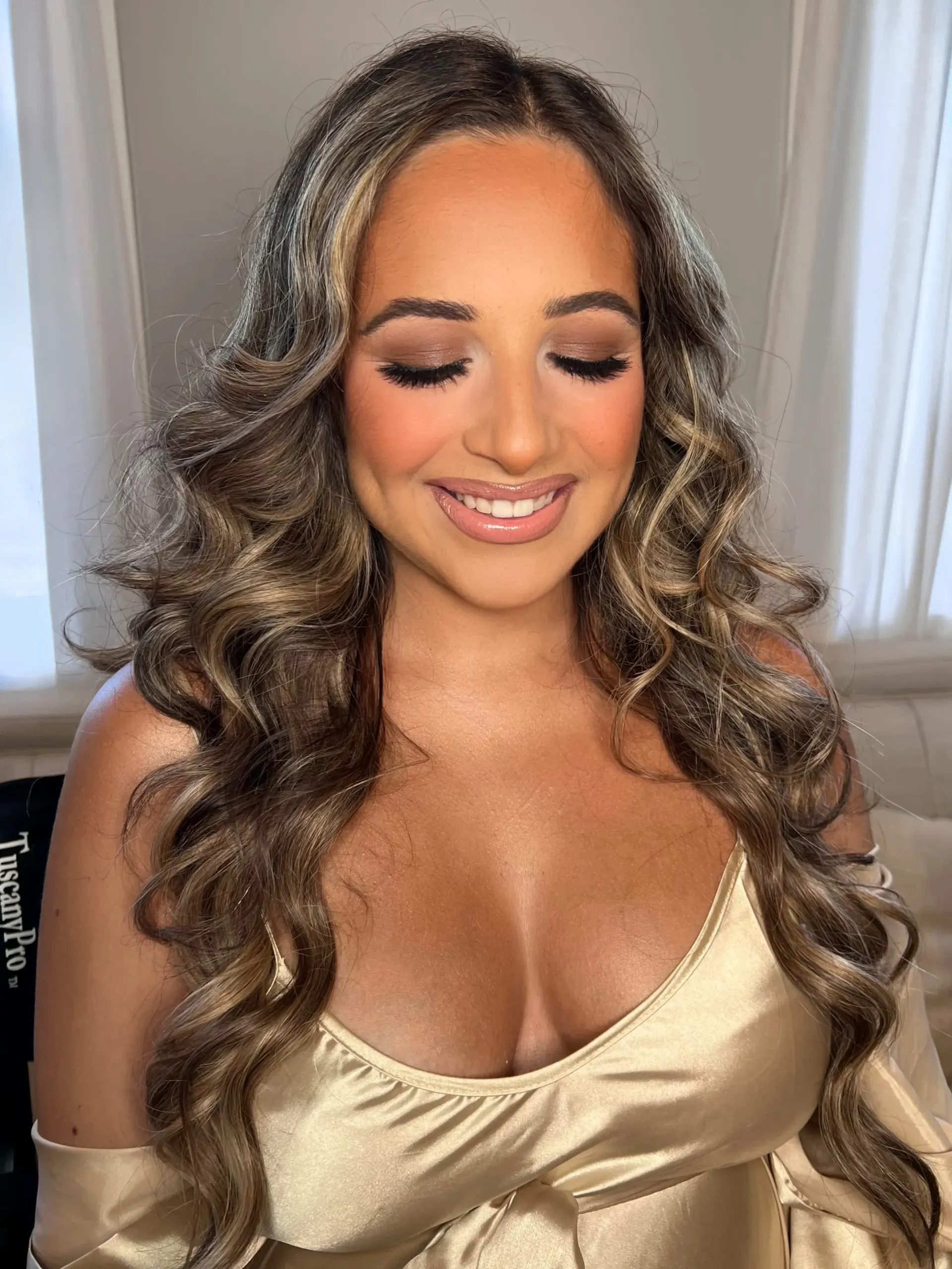 Woman with long wavy brown hair, wearing a gold dress, smiles with eyes closed, heavy makeup, indoors.