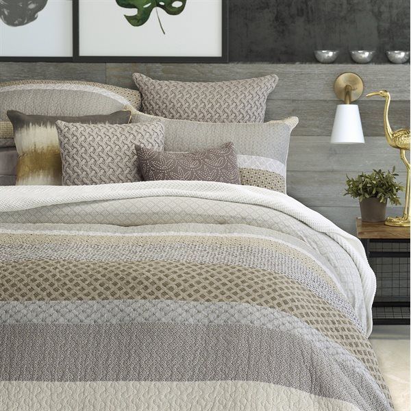 A bed layered with a neutral-toned, striped textured comforter and various pillows in a bedroom with wooden wall paneling.