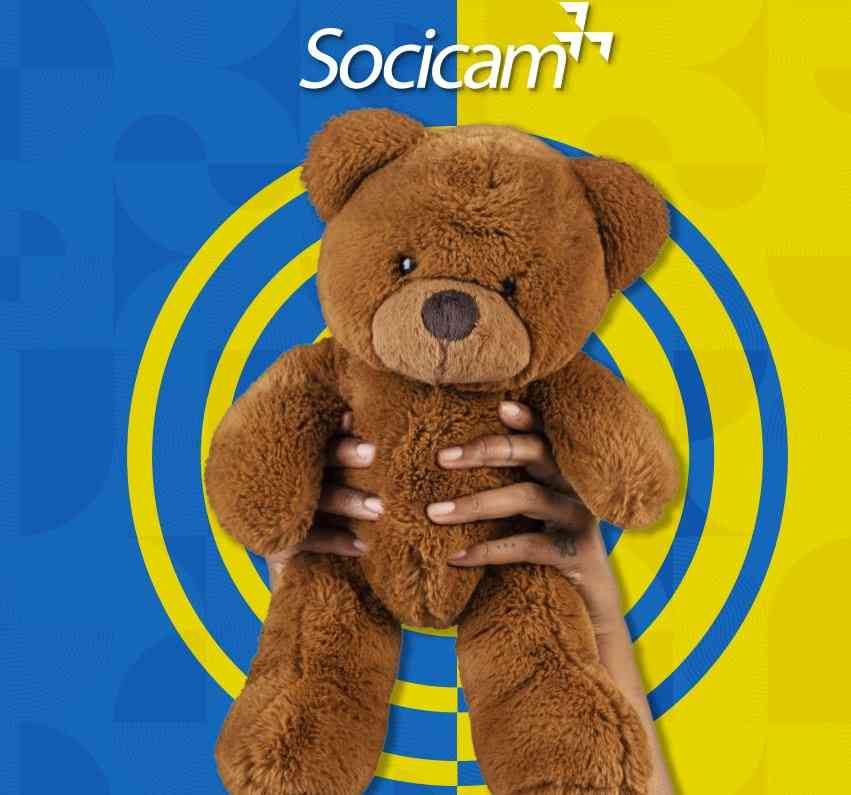 Urso de pelúcia marrom segurado por mãos, centralizado em um fundo azul e amarelo com o logotipo da Socicam.