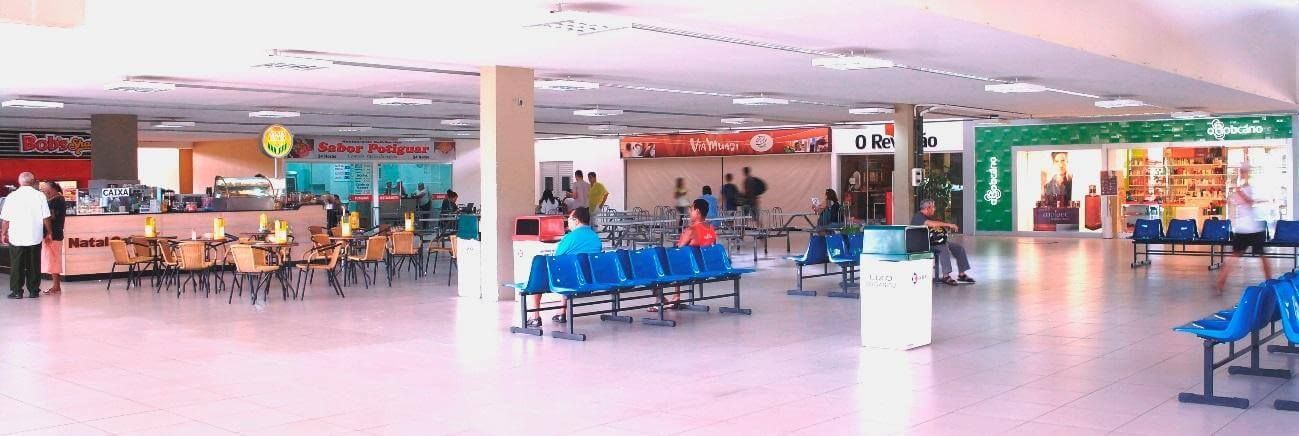 Área de espera bem iluminada do aeroporto, com fileiras de assentos azuis, balcões e algumas pessoas no interior.