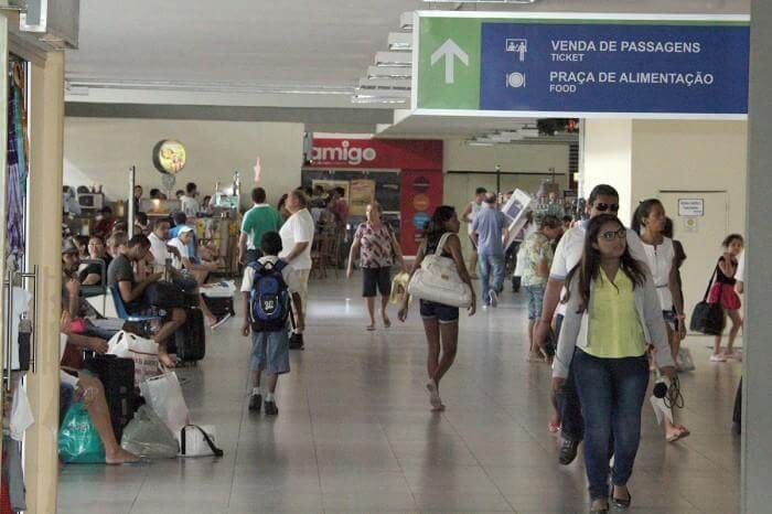 Corredor interno lotado de aeroporto, com viajantes passando por placas de alimentação e serviços.