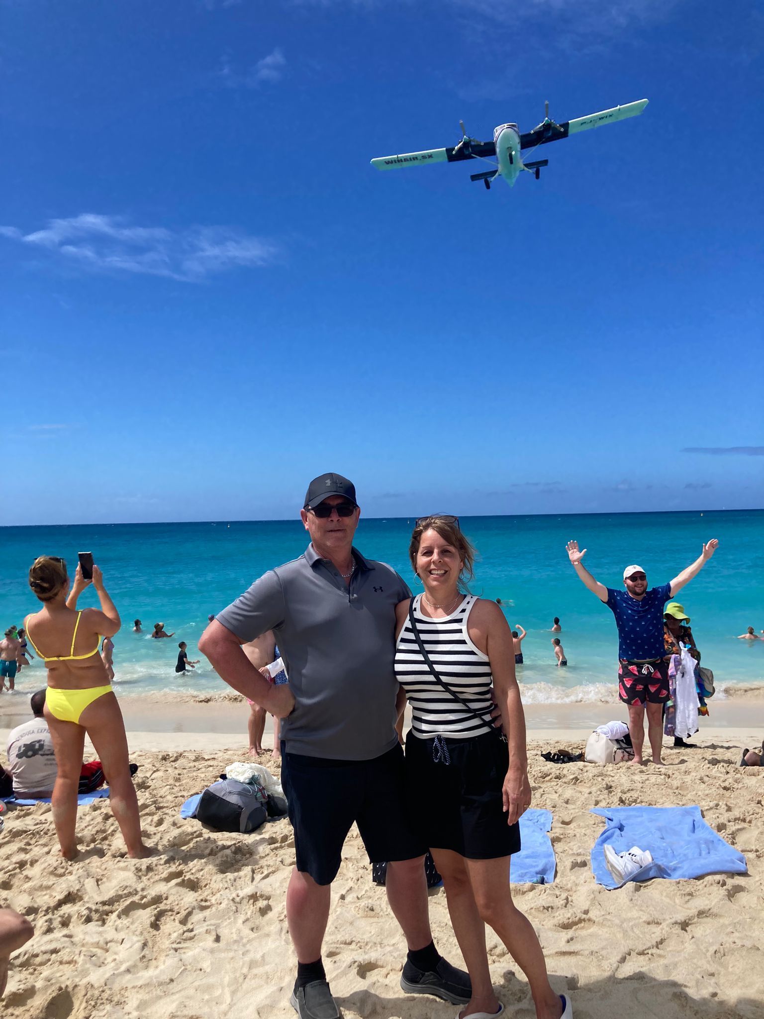 A man and a woman are standing on a beach with a plane flying overhead.