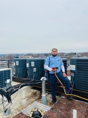 Man sitting on a rooftop HVAC unit, holding a pressure-washing wand near rooftop equipment.