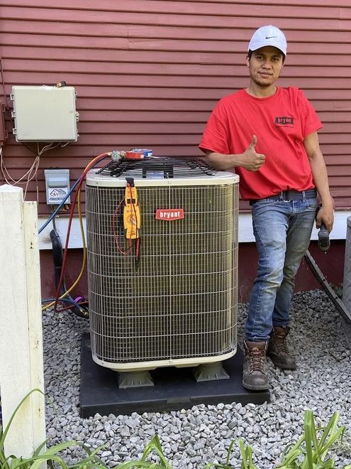 Man in red shirt standing beside outdoor HVAC unit, giving a thumbs-up by a house wall.