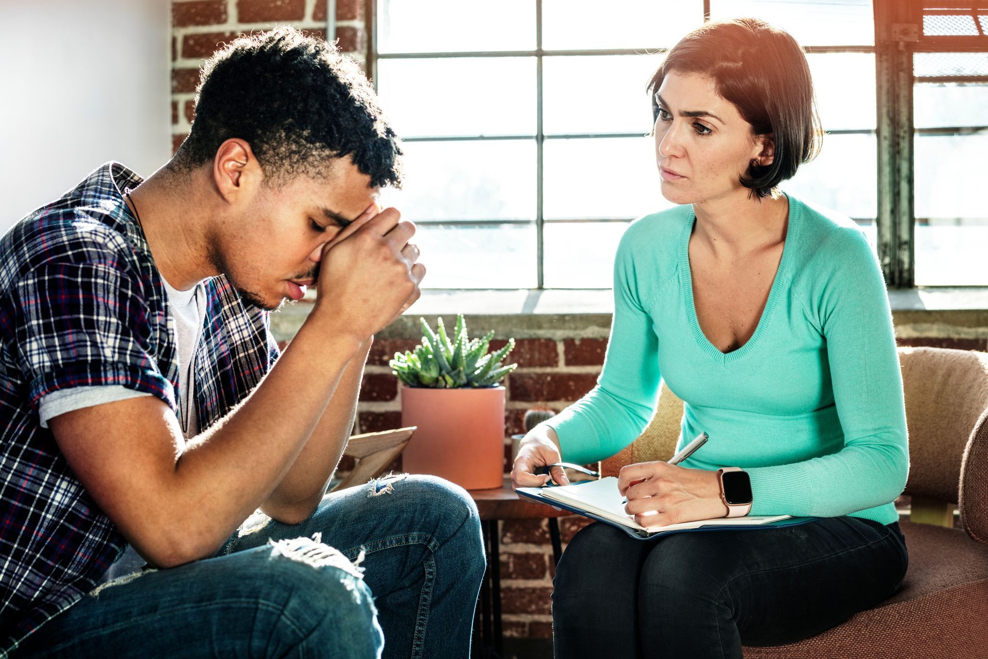 A person sits with their head in their hands while a counselor observes and takes notes in a sunny, brick-walled office.
