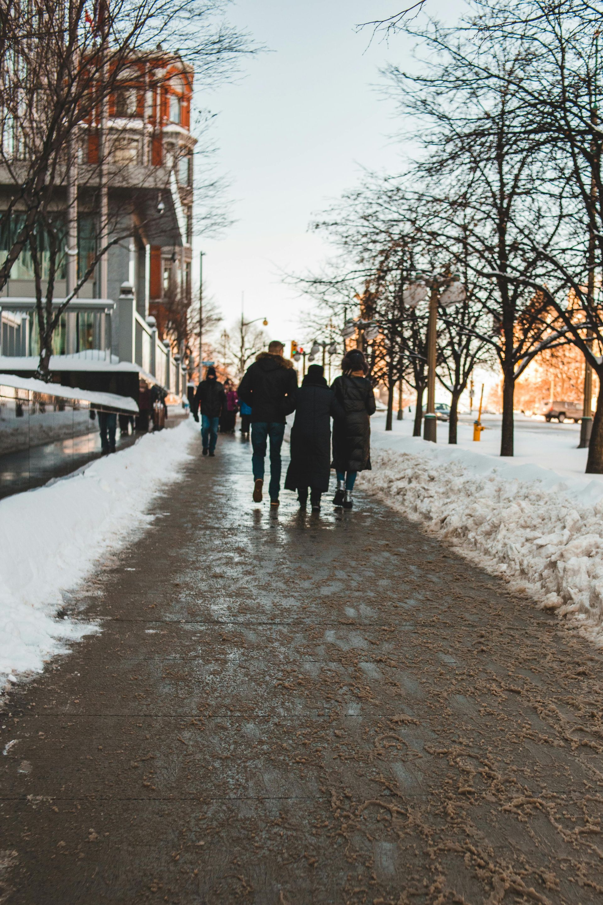 People walking on a snowy sidewalk next to a building and bare trees. Winter scene.