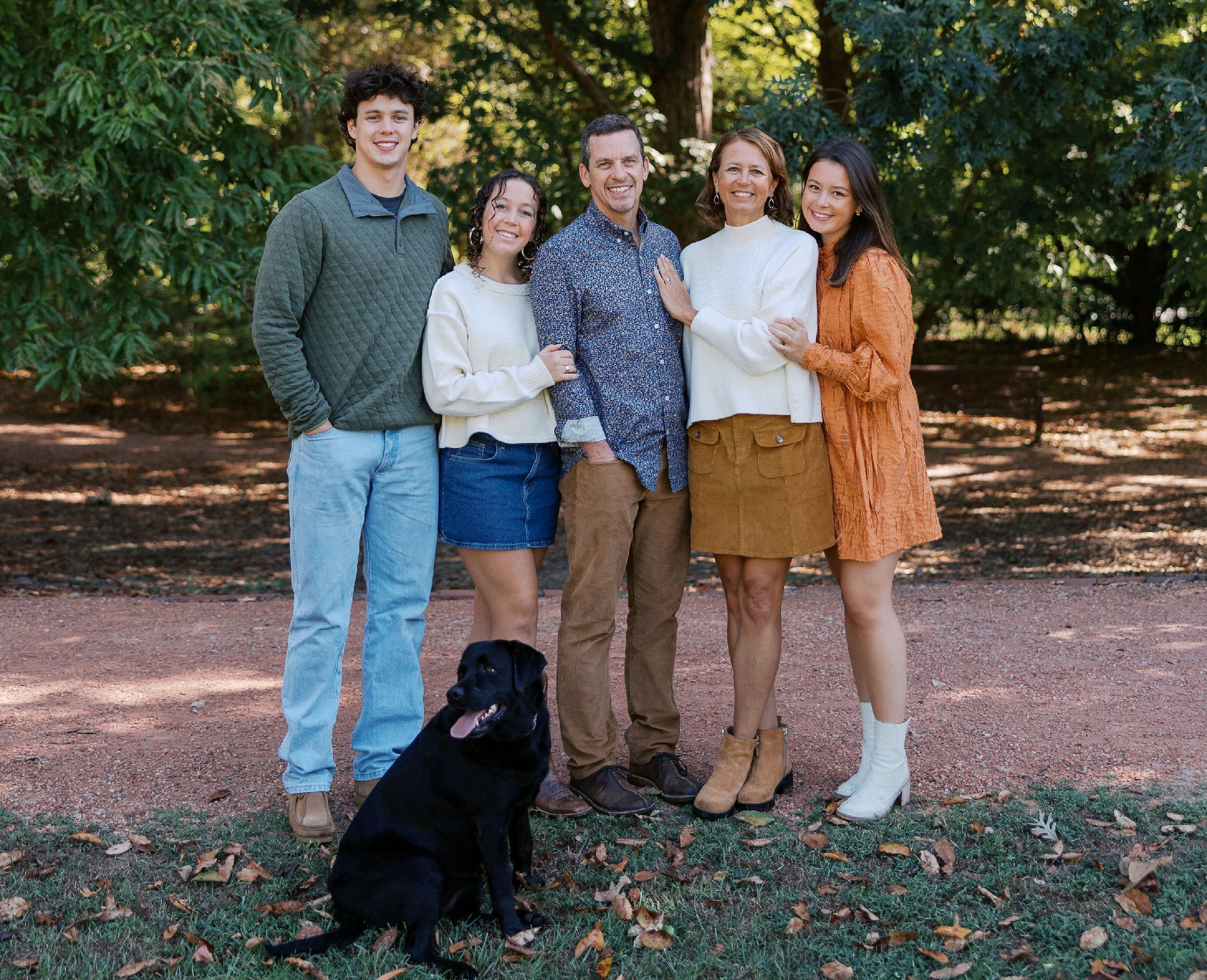 Family of five and a black dog posing outdoors. Woman in orange dress, others in sweaters.