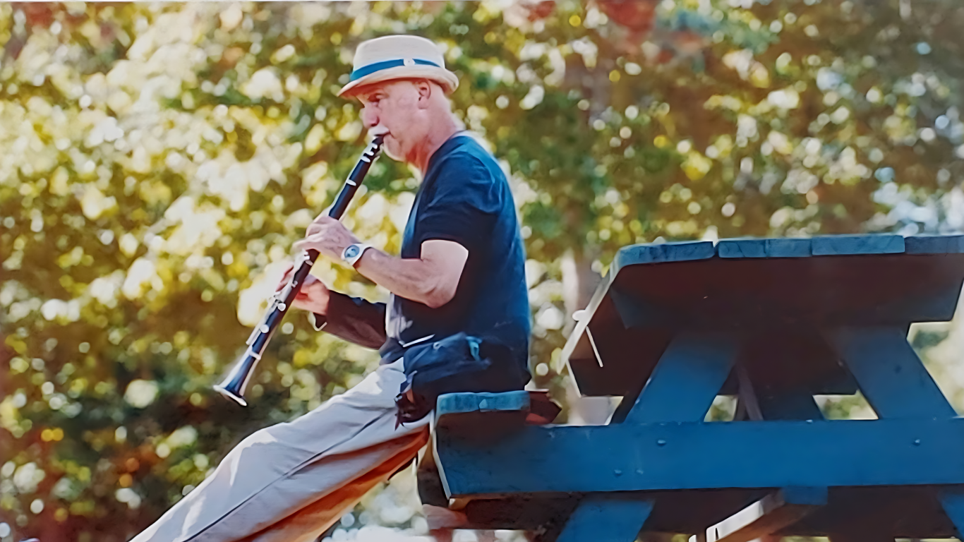 Man in a hat plays the clarinet seated on a park picnic table. Green foliage in the background.