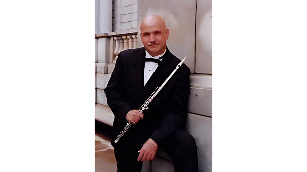 Man in tuxedo holding a flute, leaning against a stone wall.