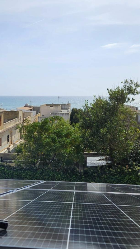 Pannelli solari su un tetto con vista sull'oceano, alberi ed edifici sotto un cielo azzurro.