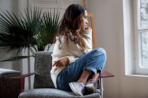 Woman sitting cross-legged on a chair by a bright window, looking outside with a calm expression.