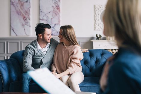 Two people sit on a couch talking in a cozy living room, with pillows and soft daylight.