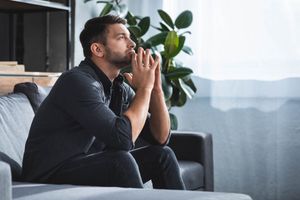 Man sitting on a couch with hands clasped, looking out a window in a softly lit living room.