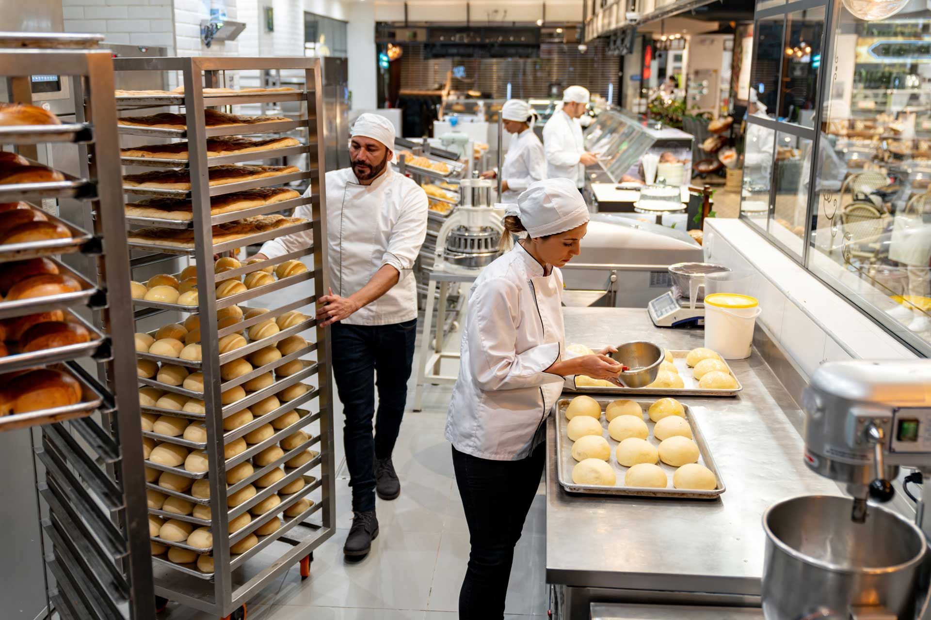 Bakers in a professional kitchen prepare trays of dough, one moving a tall rack filled with bread while another works.