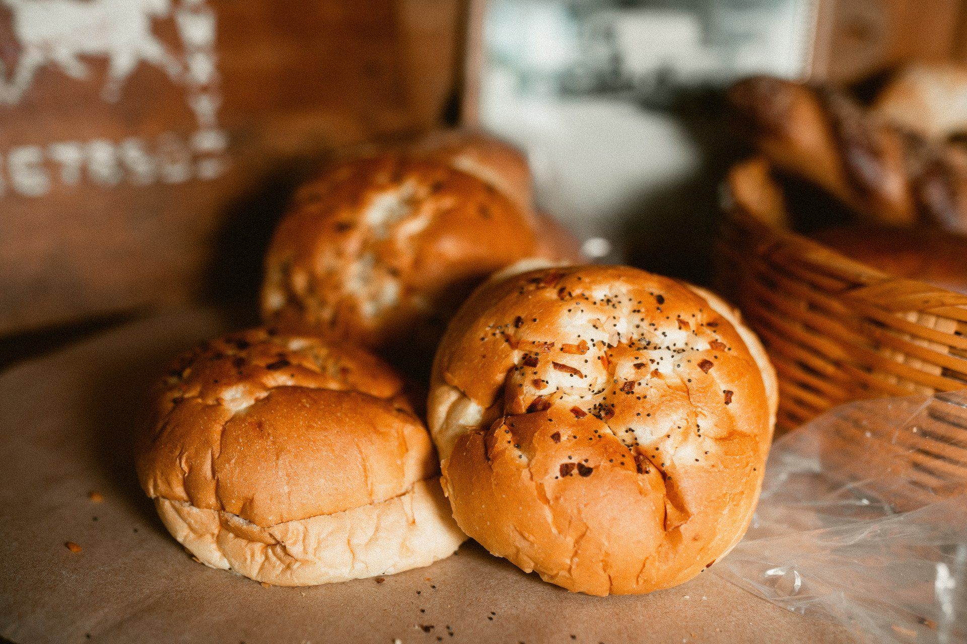 Three golden-brown dinner rolls sprinkled with herbs sit on a brown paper surface next to a wicker basket.