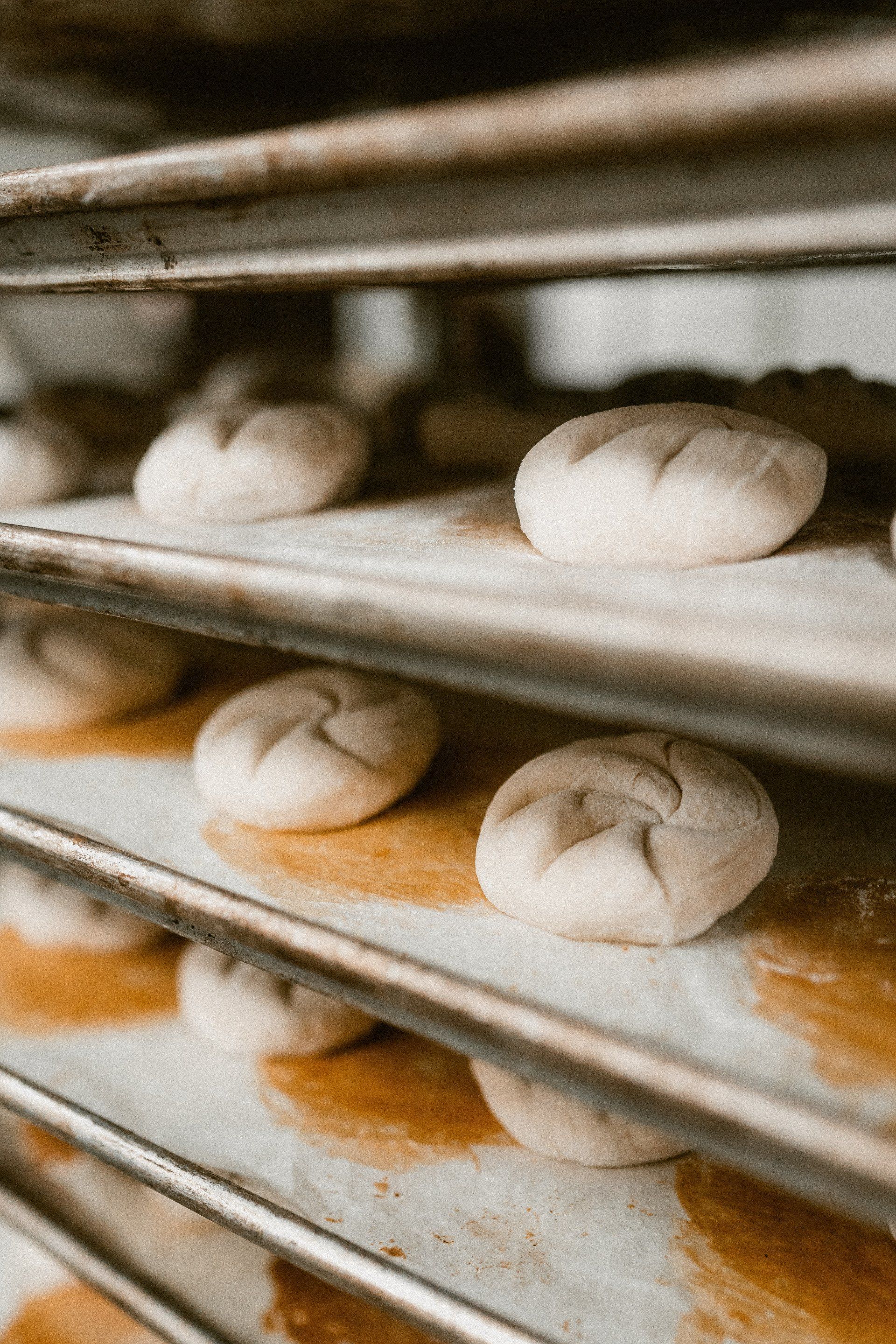 Raw, shaped bread dough rolls resting on multiple stacked metal baking sheets in a kitchen.